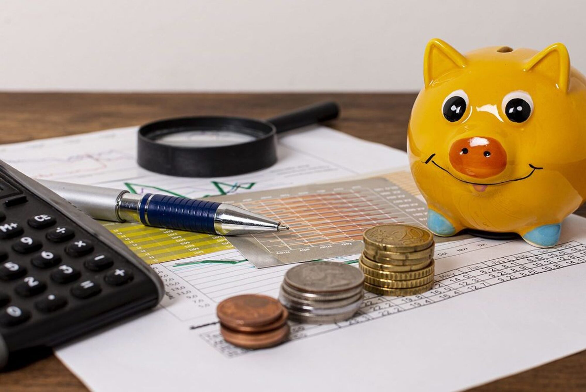 Un cochon jaune à côté de pièces de monnaie, d'une calculette, d'un stylo et d'une loupe posés sur une feuille de calcule sur une table en bois